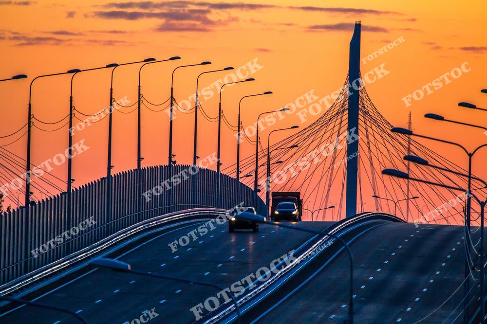 Фотография современного Санкт-Петербурга с видом на мост Кольцевой автодороги через Неву рядом с Крестовским островом. На снимке запечатлена масштабная транспортная инфраструктура северной столицы России, соединяющая различные районы города. Высокие опоры моста, широкая магистраль и водная гладь Невы создают динамичный городской пейзаж. Панорамный вид подчеркивает сочетание современной архитектуры, инженерных сооружений и природных видов Финского залива. Изображение отлично подходит для туристических публикаций, архитектурных проектов, интерьерных постеров, сувенирной продукции и коллекций фотографий Санкт-Петербурга.